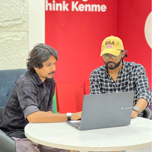 Digital Marketing Strategist in Malappuram - Two people are seated at a round table in front of a laptop. One wears a yellow cap and glasses, and the other has short hair. They appear to be collaborating on something. A red and white wall with partial text is behind them.