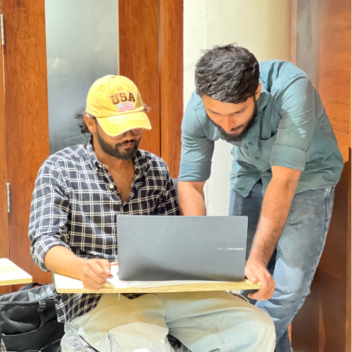 Digital Marketing Strategist in Malappuram - Two people are collaborating over a laptop. One is sitting, wearing a checkered shirt and yellow cap, while the other stands beside him, in a teal shirt, leaning in to look at the screen. They are indoors, with wood paneling in the background.