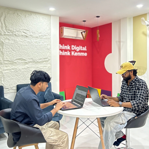 Digital Marketing Strategist in Malappuram - Two people are working on laptops at a round table in a modern office space. The wall behind them features bold text and vibrant colors. They are seated on black chairs, focused on their screens.