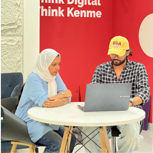 Digital Marketing Strategist in Malappuram -Two people sitting at a round table are working on a laptop. The woman wears a hijab, and the man wears a cap. A red banner on the wall reads "Think Digital Think Kenme.