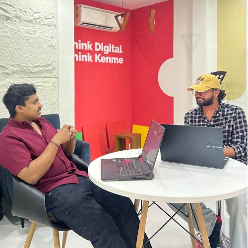 Digital Marketing Strategist in Malappuram -Two people seated at a round table in a modern office setting, each with an open laptop. One is wearing a red shirt and the other a black checkered shirt with a cap. The wall behind them reads "Think Digital Think Kenme" in bold letters.