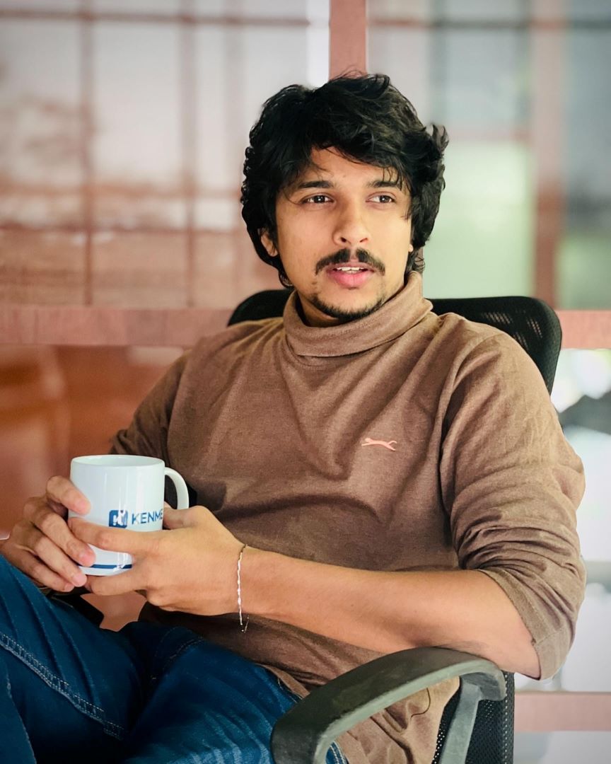 Digital Marketing Strategist in Malappuram- A person with wavy hair sits in a chair, holding a white mug. They're wearing a brown long-sleeve shirt and jeans, gazing thoughtfully to the side. The background features glass panels.