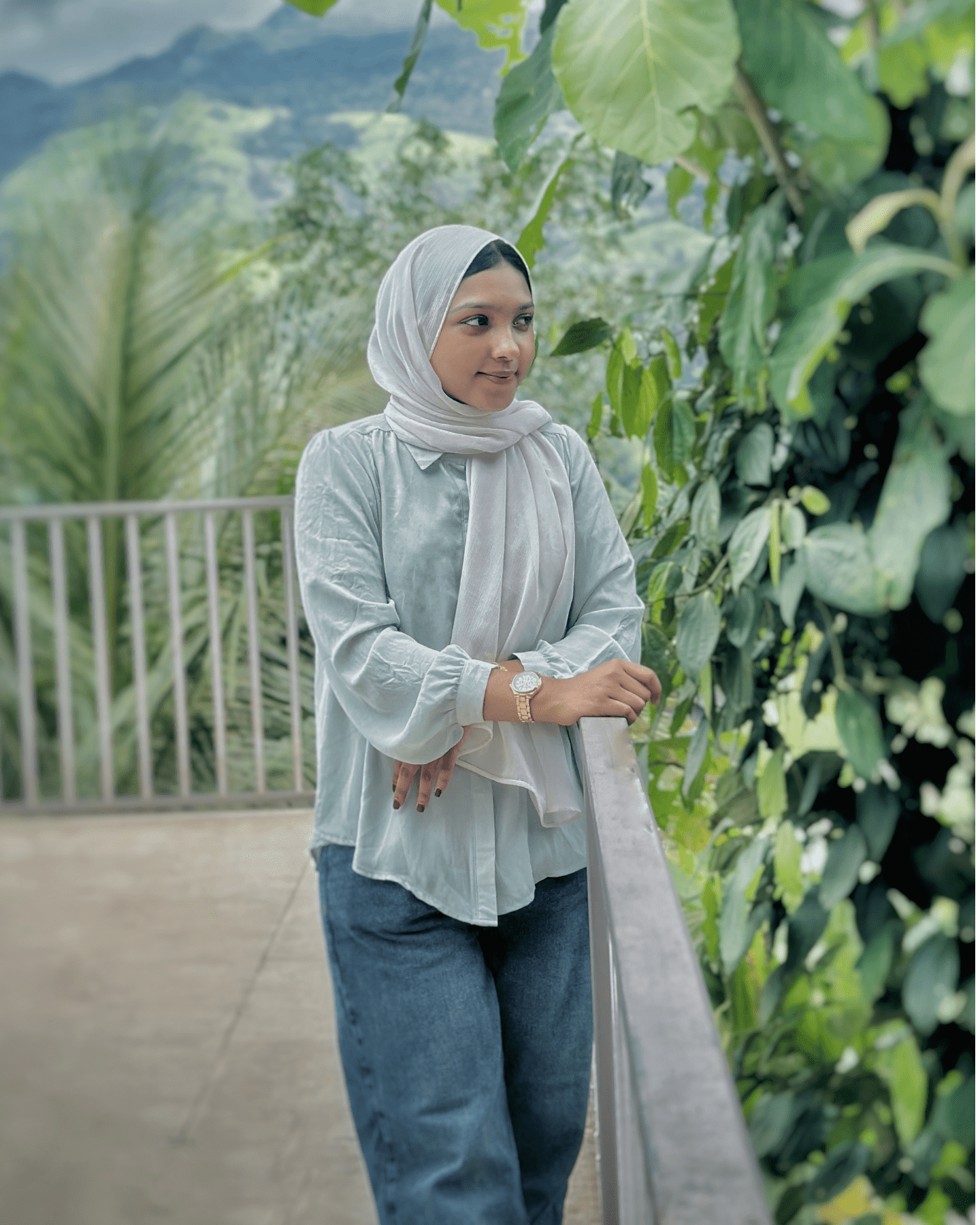 Digital Marketing Strategist in Malappuram - A woman in a light-colored hijab and shirt leans on a railing, surrounded by lush greenery. The background features blurred mountains under a cloudy sky.