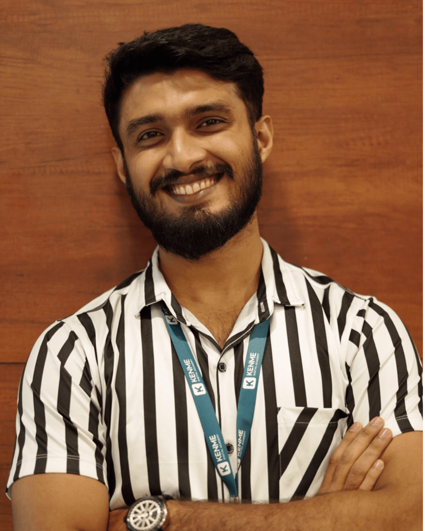 Digital Marketing Strategist in Malappuram - A man with a beard is smiling, wearing a black and white striped shirt and a lanyard, standing in front of a wooden wall.