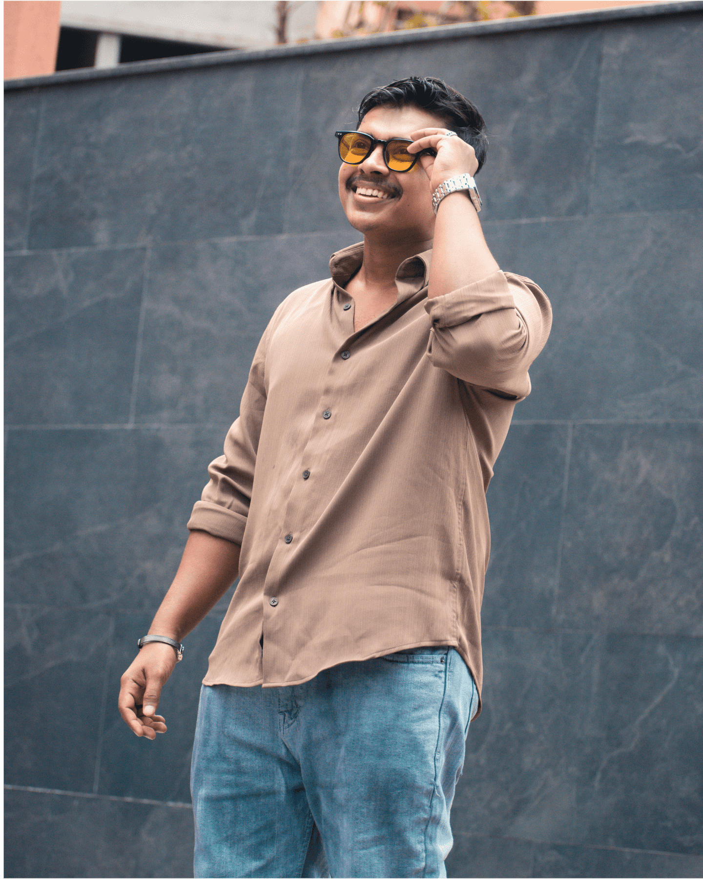 Digital Marketing Strategist in Malappuram - A smiling person wearing sunglasses, a beige shirt, and jeans stands against a dark textured wall. They have short hair and are touching their sunglasses with one hand.