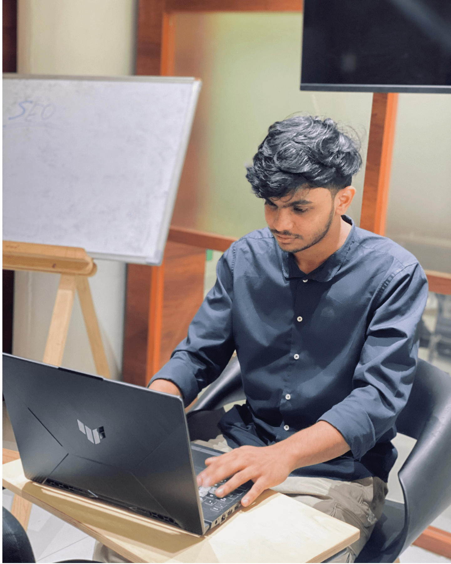 Digital Marketing Strategist in Malappuram - A person in a black shirt is sitting on a chair, typing on a laptop at a small table. In the background, there's a whiteboard and a wooden easel.