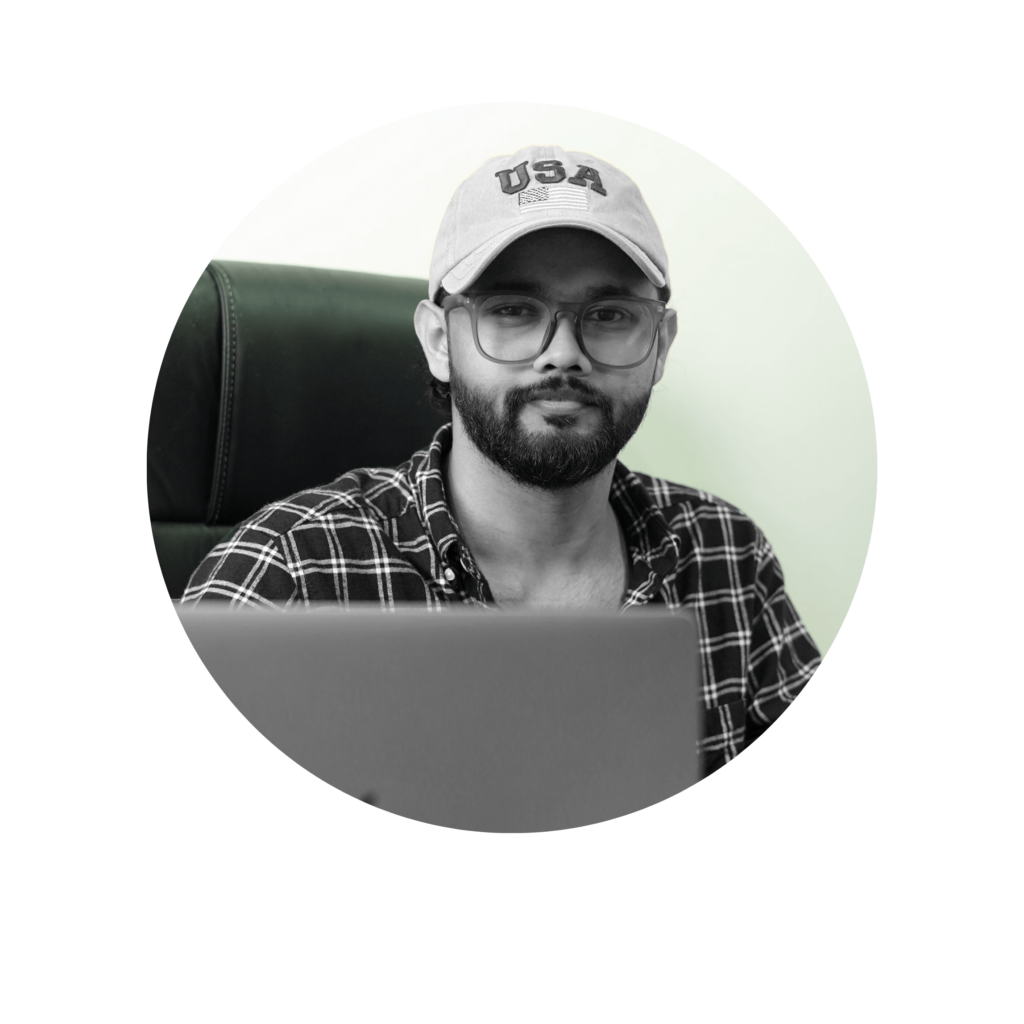 Digital Marketing Strategist in Malappuram - A person with a beard and glasses sits in front of a laptop. They are wearing a checked shirt and a cap with "USA" printed on it. The background is minimal, with a chair visible. The image is in black and white.